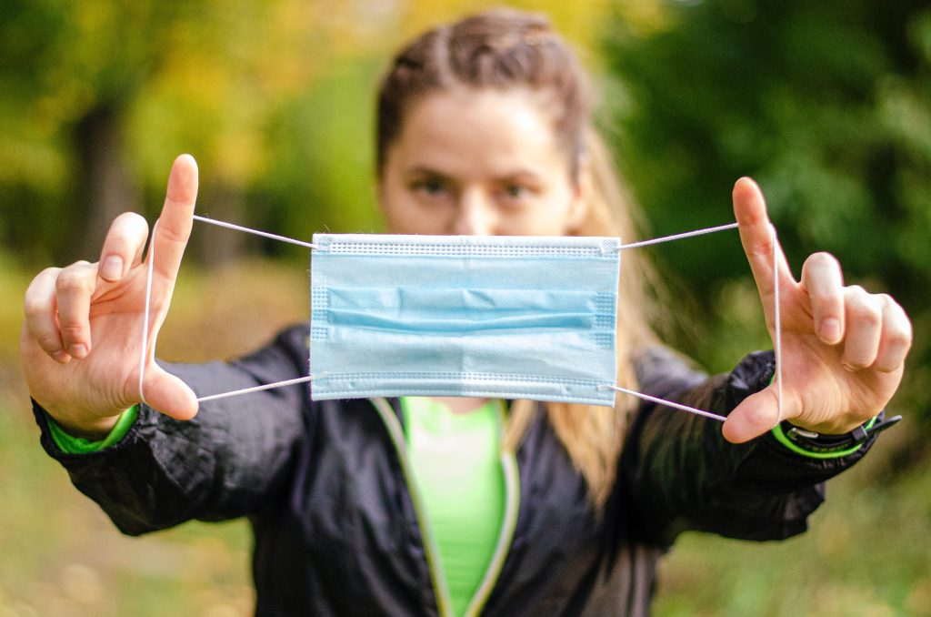 Environmental Impact of PPE - Caucasian blonde women in the background holding out a PPE disposable mask stretched between her hands placed in front of her at shoulder length - Cait Bagby