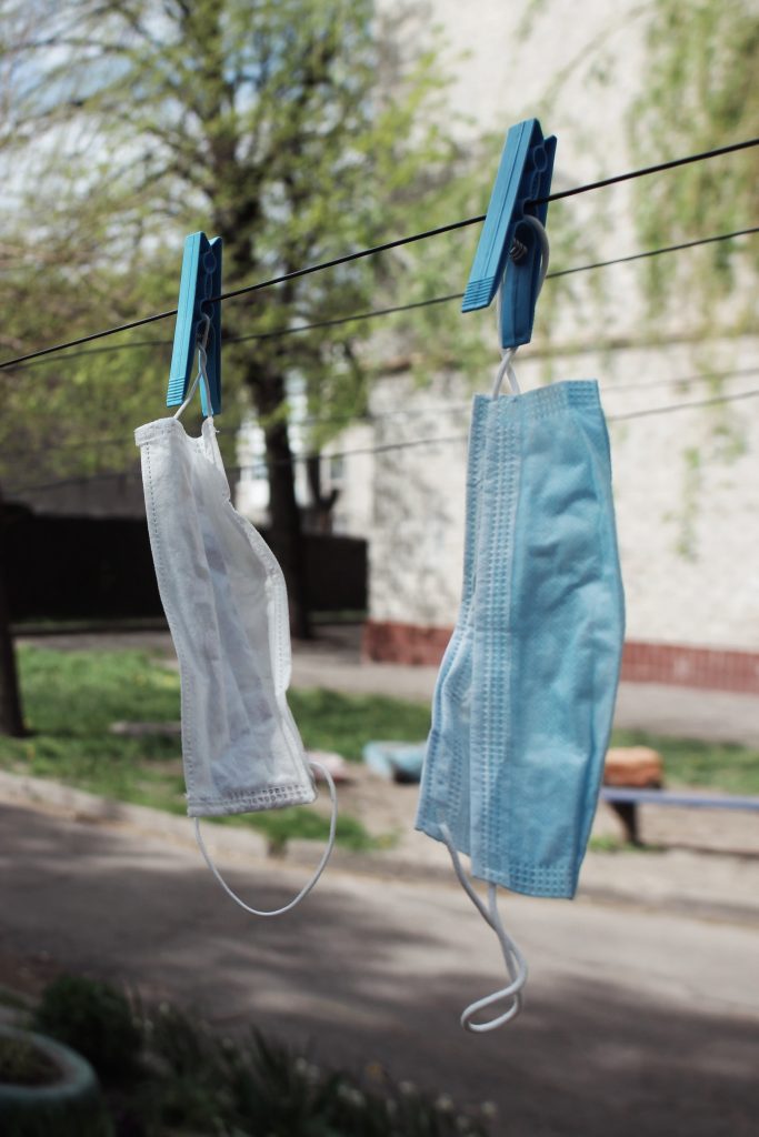 Environmental Impact of PPE - Photo of one white and one blue disposable PPE masks hanging on a laundry line being kept in place with blue laundry clips - Cait Bagby