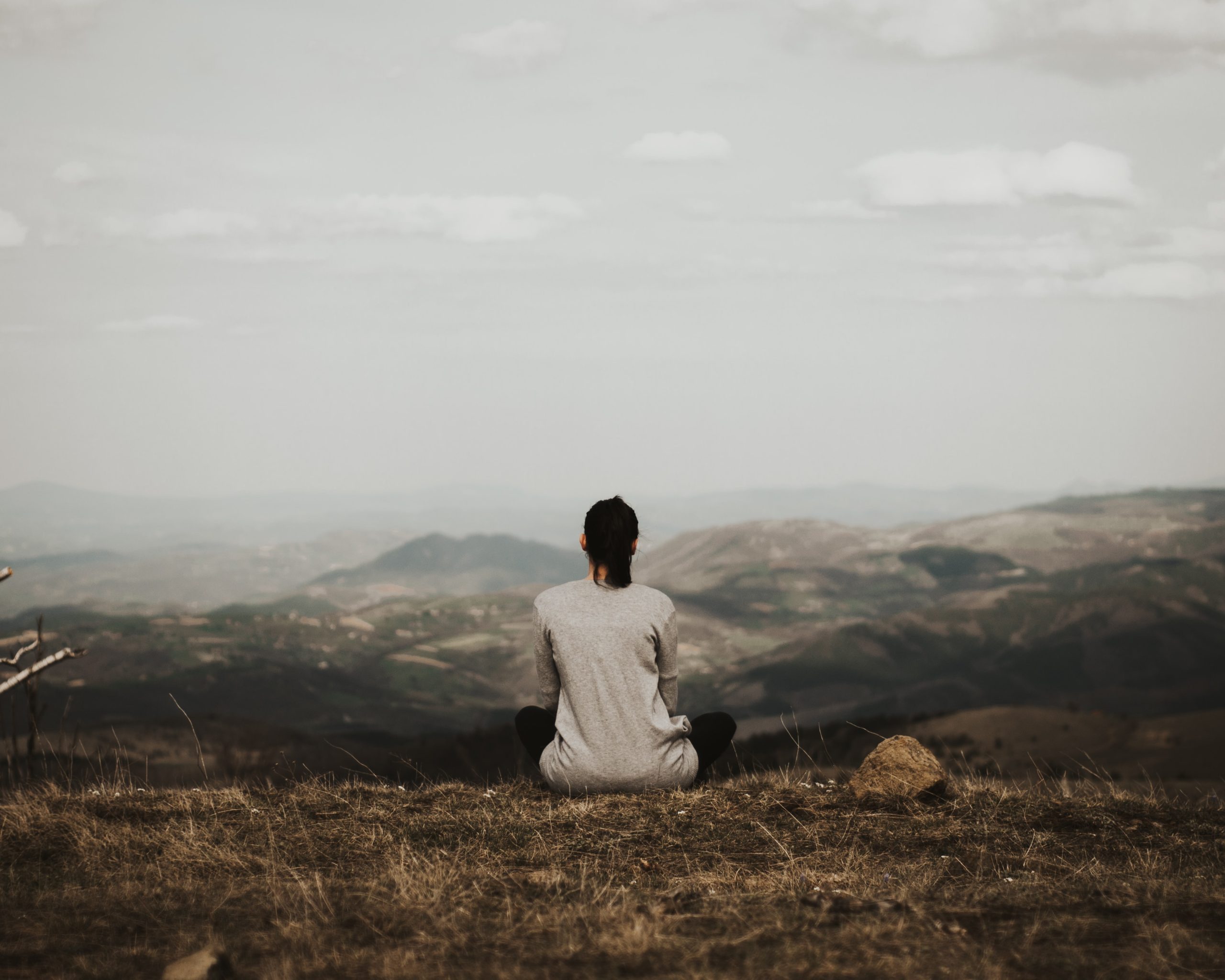 Caucasian woman in a white long tshirt with dark hair is sitting on a brown tinged hill looking at mountains in the distance. Article about empathy for the earth