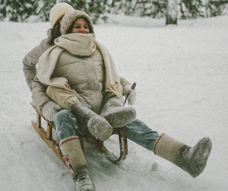 Low Cost, Low Waste Valentine's Day Ideas World Threads Traveler, Cait Bagby. Two Women wearing off white puffer coats, beige boots, white scarves and hats sledding together on a wooden sled in a snowy forest