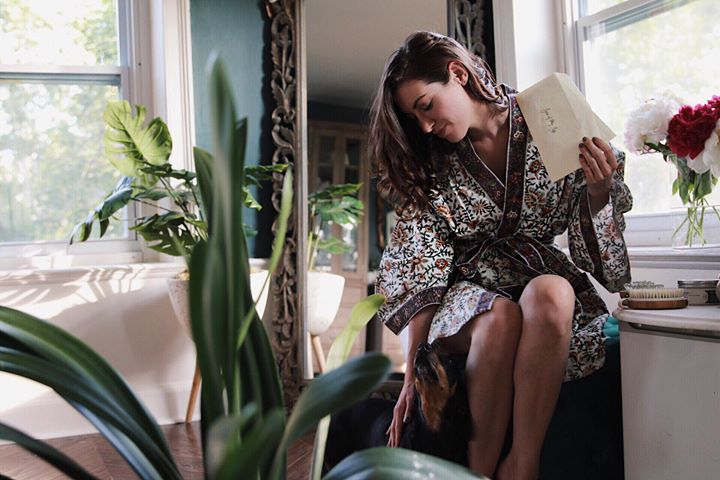 Low Cost, Low Waste Valentine's Day Ideas World Threads Traveler, Cait Bagby in a Fair Trade paisley robe sitting on a green bench in a bathroom with a full length mirror behind her holding a handwritten love letter.
