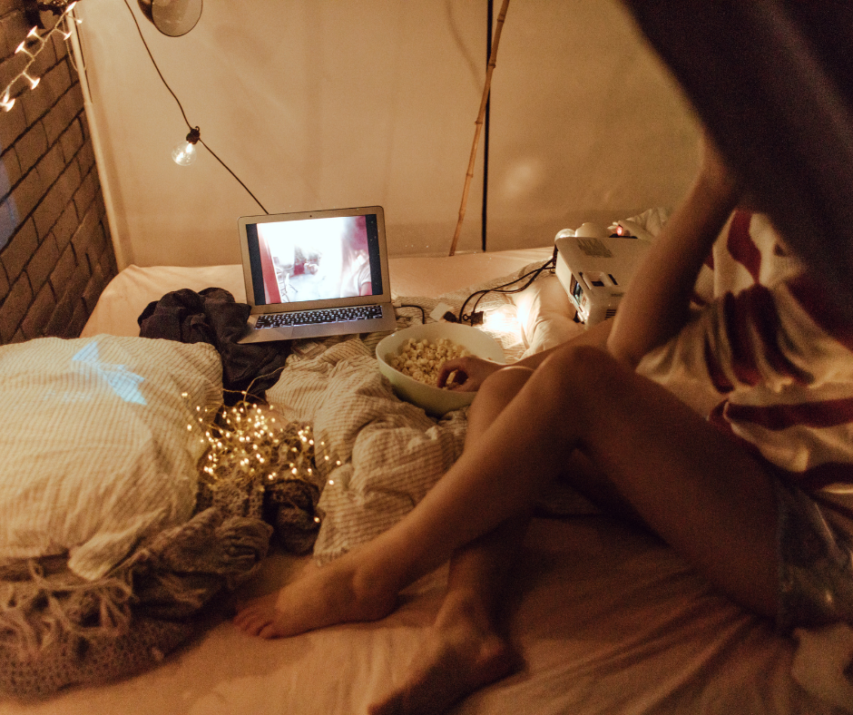 Low Cost, Low Waste Valentine's Day Ideas World Threads Traveler, Cait Bagby a woman sitting on an unmade bed with white and beige sheets and fairy lights in the background watching a movie on her computer.