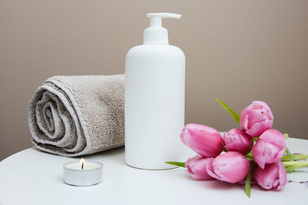 Low Cost, Low Waste Valentine's Day Ideas World Threads Traveler, Cait Bagby - photo of a beige towel rolled up next to a white pump bottle, pink tulips and a tea light on a white table - home spa day
