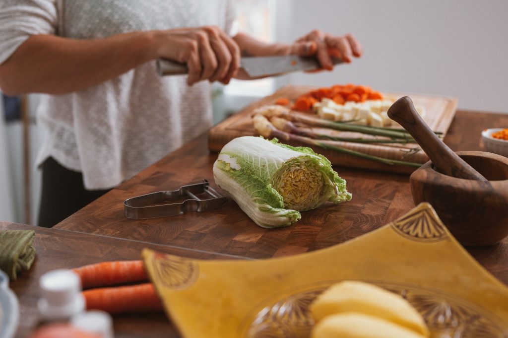 Low Cost, Low Waste Valentine's Day Ideas World Threads Traveler, Cait Bagby, Image of a woman holding a knife above a wooden cutting board with chopped onions, carrots and lettuce on the board.