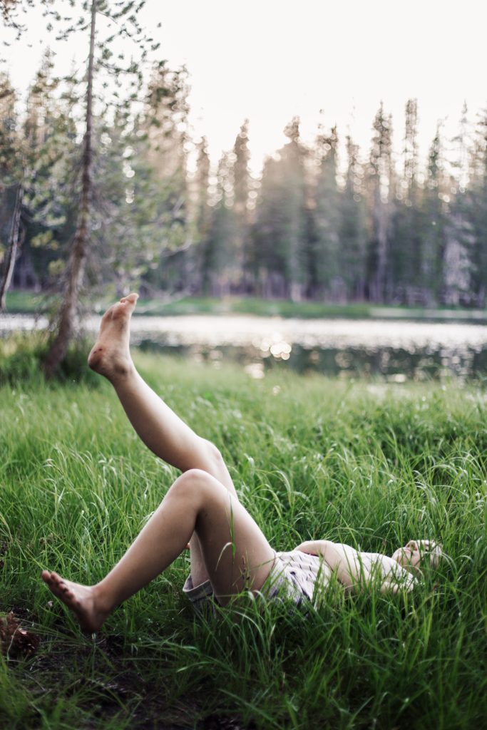 Benefits of Connecting with Nature - World Threads Traveler. Young caucasian individual laying in tall green grass with pine trees and a pond in the background. One foot is up in the air. They are connecting with nature